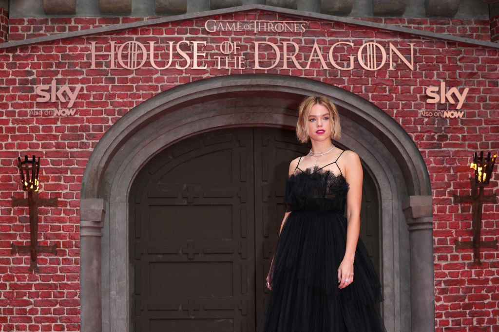 Milly Alcock in a black dress in front of a brick wall. 